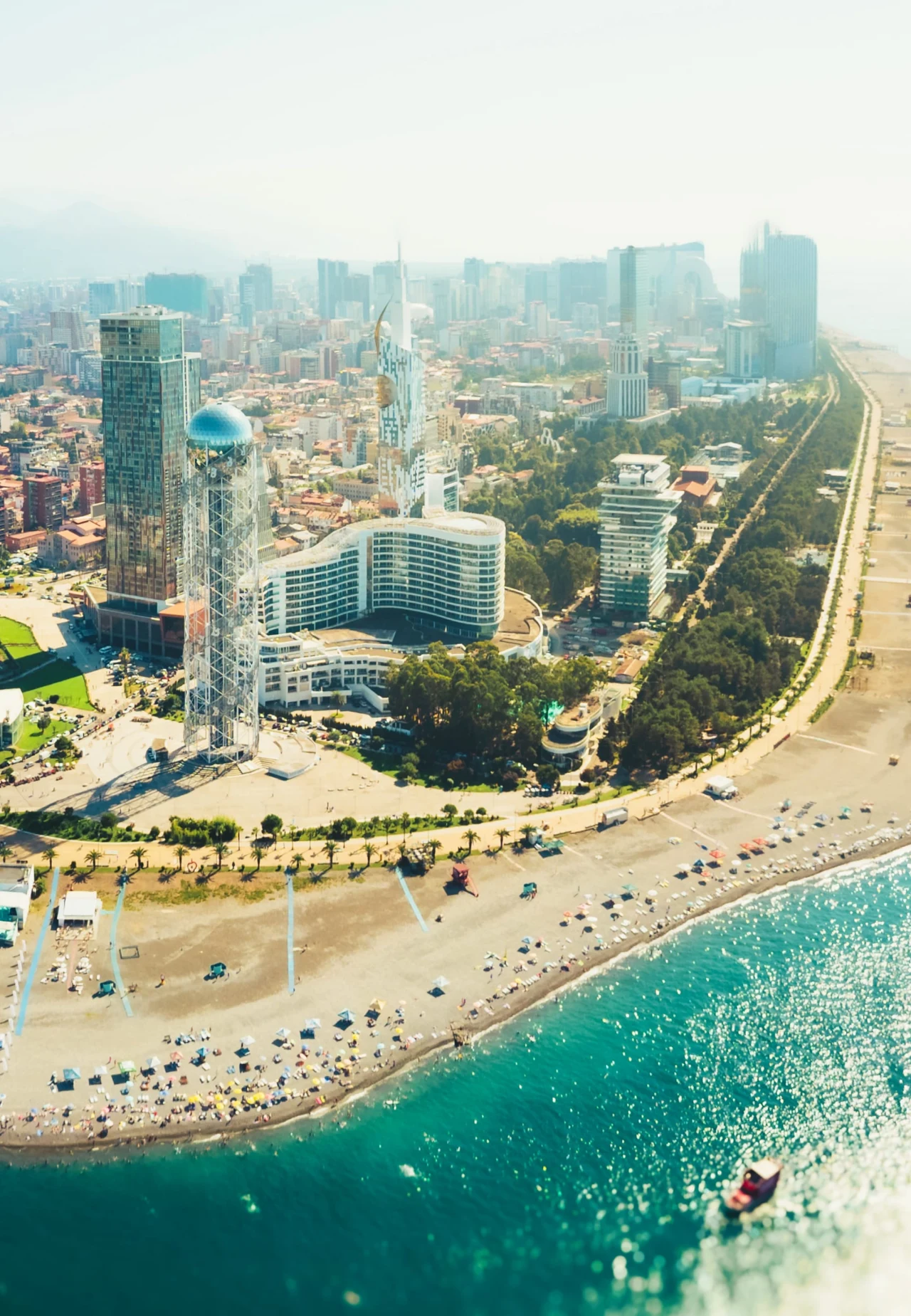 Nowoczesne wieżowce i charakterystyczne budynki przy szerokiej, piaszczystej plaży, przy której rozstawione są parasole i leżaki, a w pobliżu brzegu widać łódź na spokojnej, błękitnej wodzie.
