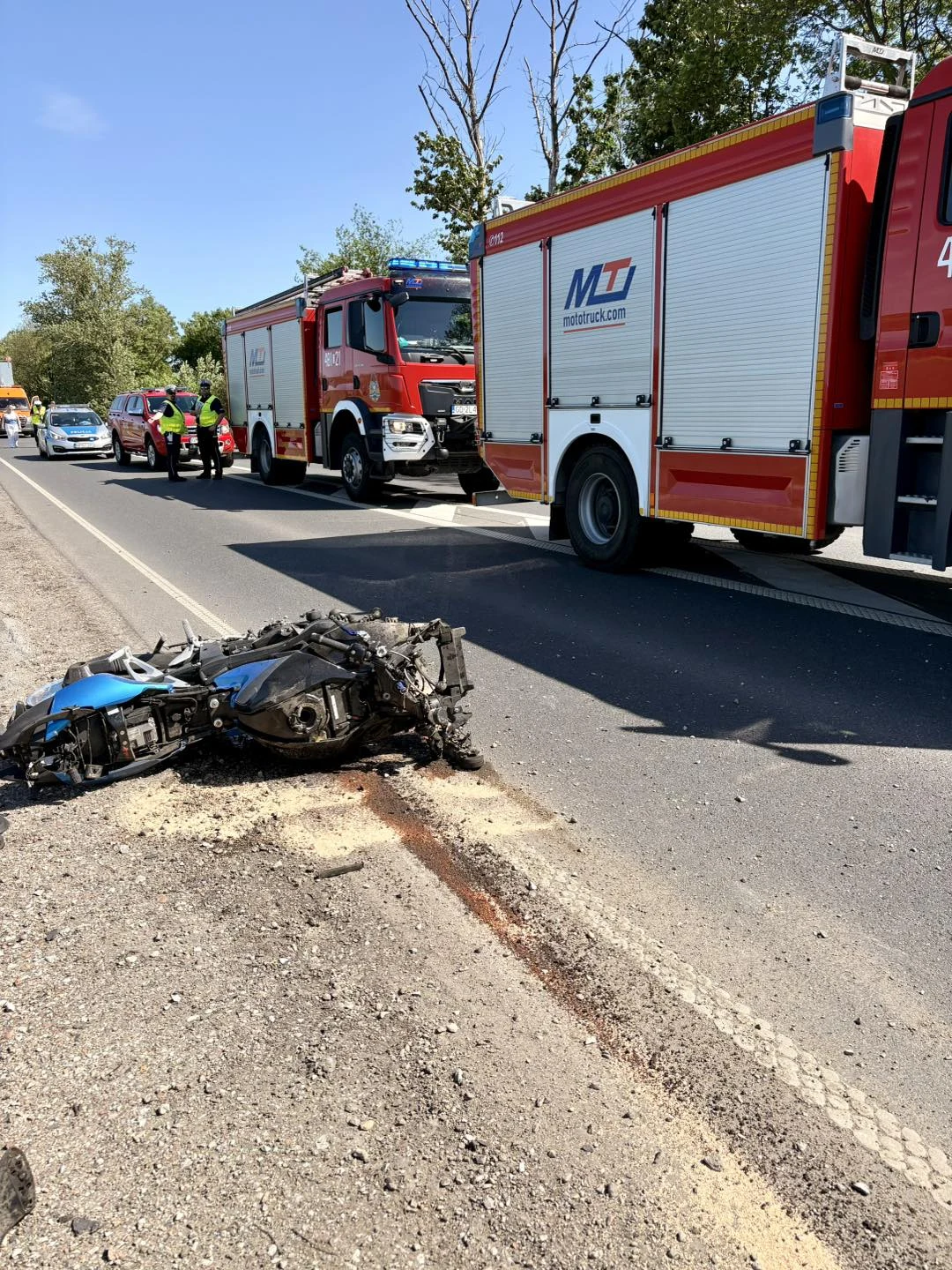 Rozbity motocykl leży na poboczu asfaltowej drogi, a przy nim znajduje się ślad płynów oraz piasku; na jezdni stoją dwa wozy strażackie, obok których pracują strażacy w odblaskowych kamizelkach.