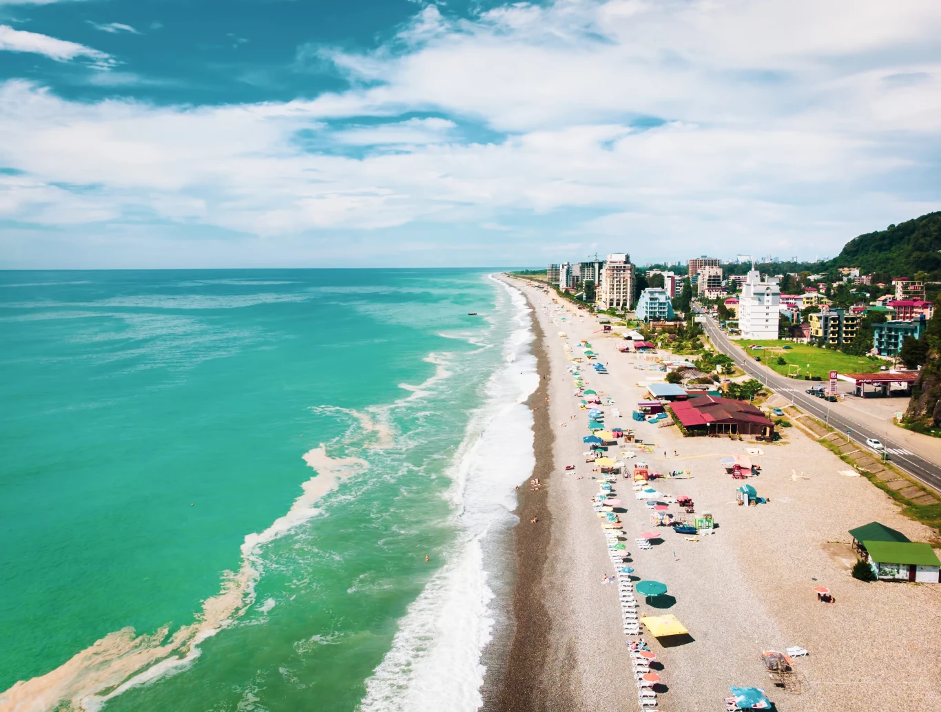 Szeroka, kamienista plaża z kolorowymi leżakami i parasolami wzdłuż brzegu, turkusowe morze po lewej, po prawej linia hoteli i budynków oraz zielone wzgórze w tle.