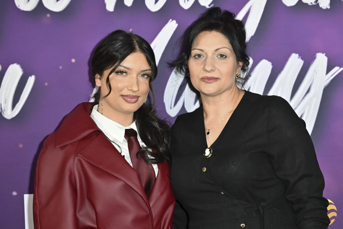 Dwie kobiety pozujące razem na tle fioletowej ścianki z białymi napisami, jedna w bordowej marynarce i białej koszuli, druga w czarnej bluzce z dekoltem i guzikami.