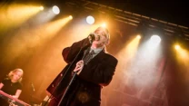 Wokalistka w czarnym garniturze śpiewa do mikrofonu na scenie oświetlonej ciepłymi światłami, a w tle widoczna jest klawiszowiec grająca na instrumentach klawiszowych.
