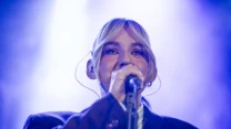 Kobieta z blond włosami spiętymi z przodu grzywką, ubrana w ciemną marynarkę, śpiewa do mikrofonu na scenie oświetlonej niebieskim światłem.