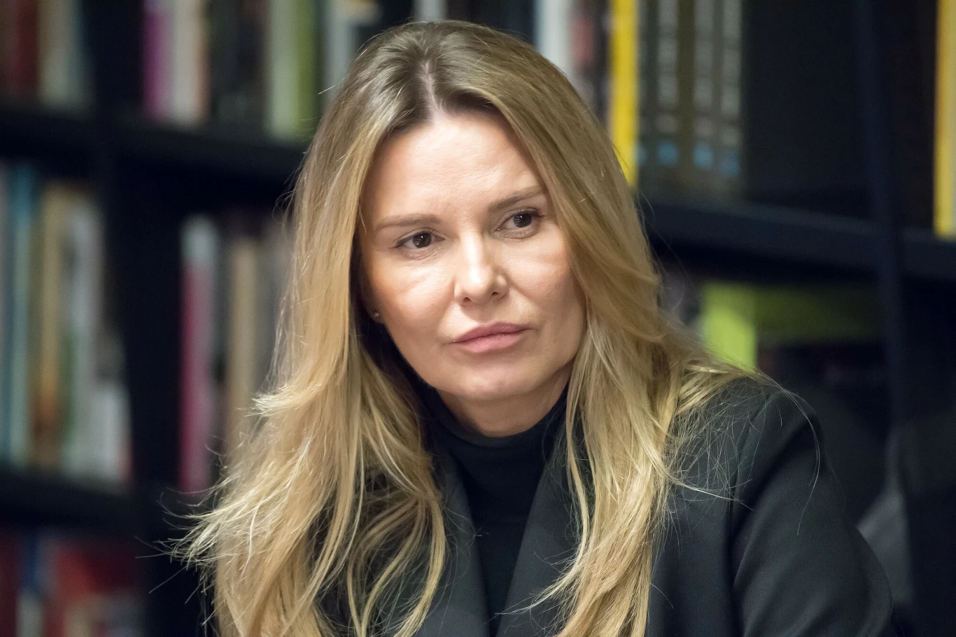 Hanna Lis Hanna Lis w czarnej marynarce siedząca przed regałem z książkami.