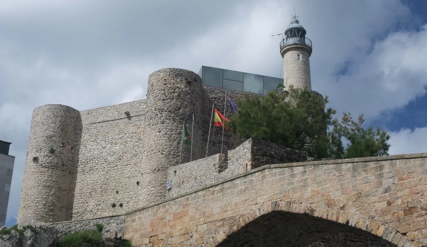 Zamek w Castro Urdiales z własną latarnią morską Zamek w Castro Urdiales z kamiennymi bastionami, trzema flagami i latarnią morską na tle nieba z chmurami.