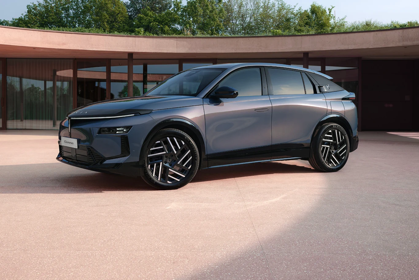 Nowoczesny SUV o futurystycznym designie z ciemnymi szybami i dużymi, geometrycznymi felgami, ustawiony na minimalistycznym dziedzińcu otoczonym przeszklonymi budynkami.
