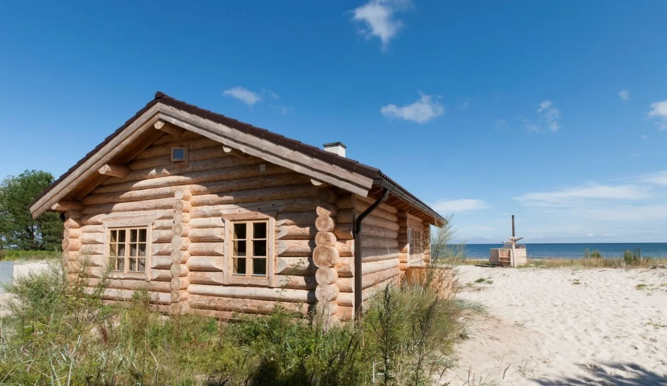 Wyspa Ruhnu zachwyca klimatem, spokojem i pustymi plażami Drewniany domek z bali przy piaszczystej plaży na wyspie Ruhnu, widok na morze i błękitne niebo.