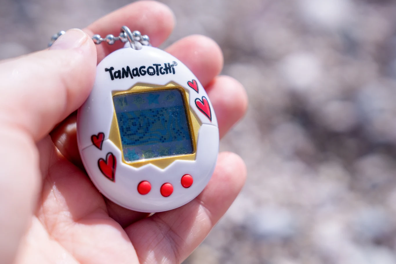 Białe Tamagotchi z czerwonymi serduszkami i trzema czerwonymi przyciskami trzymane w dłoni, z rozmazanym tłem w neutralnych barwach.