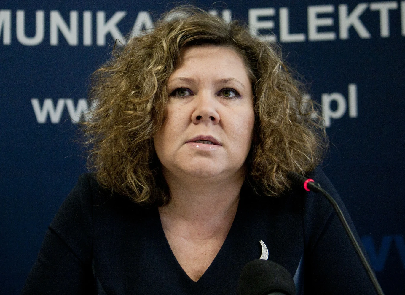 Kobieta z kręconymi włosami siedząca przy mikrofonie na tle tablicy z napisem i logotypami.