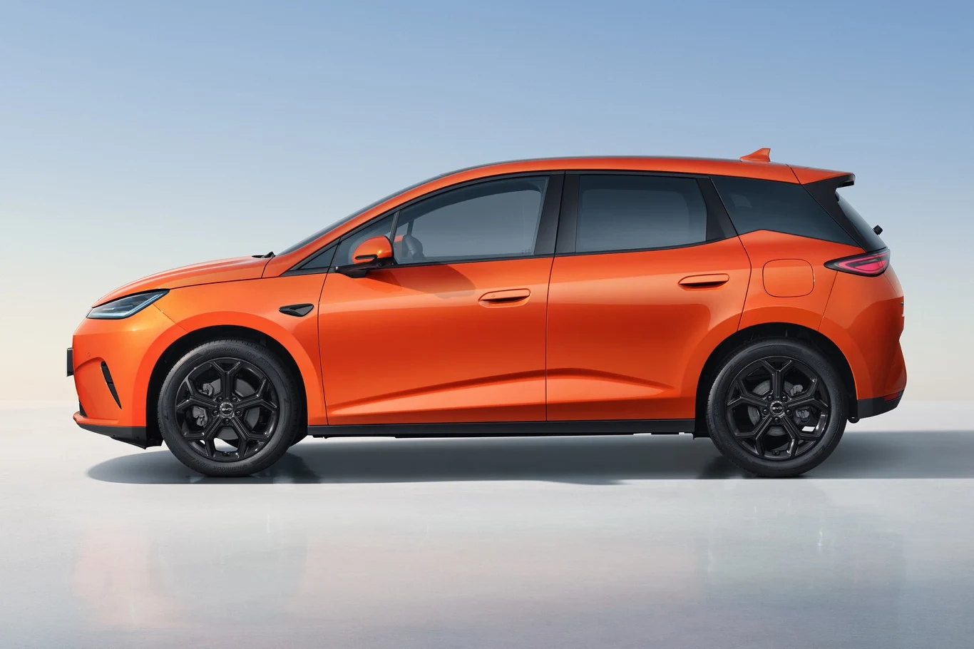 Pomarańczowy samochód typu hatchback o nowoczesnym designie, czarne felgi, przyciemnione szyby i sportowe linie nadwozia na jasnym jednolitym tle.