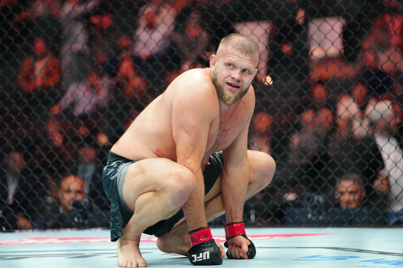 Zawodnik MMA w czarnych spodenkach i rękawicach w klatce oktagonu w pozycji kucającej, skoncentrowany i gotowy do podjęcia dalszej walki.