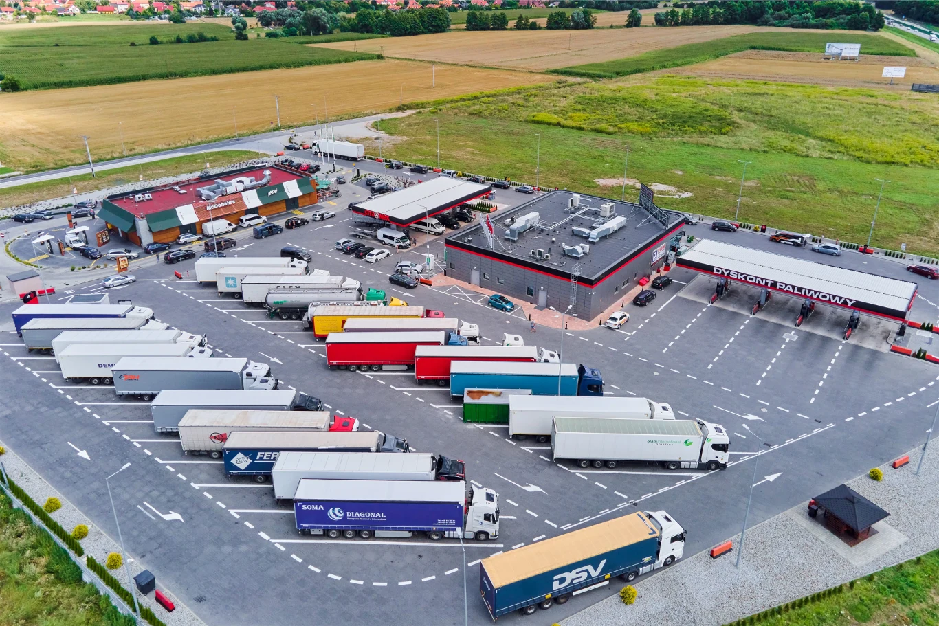 Duży parking dla ciężarówek przed stacją paliw i budynkiem usługowym, z licznymi kolorowymi naczepami ustawionymi w równych rzędach, otoczony terenami zielonymi i polami uprawnymi.