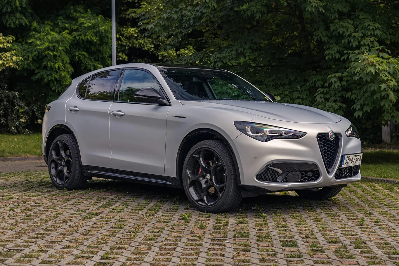 Srebrny SUV Alfa Romeo o sportowym wyglądzie stoi zaparkowany na brukowanym placu w otoczeniu zieleni.