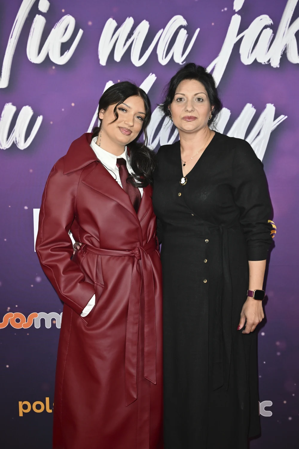 Dwie kobiety stoją obok siebie na tle fioletowej ścianki z białymi napisami; jedna z nich ma na sobie długi, bordowy płaszcz i białą koszulę z krawatem, druga ubrana jest w czarną sukienkę zapinaną na guziki.