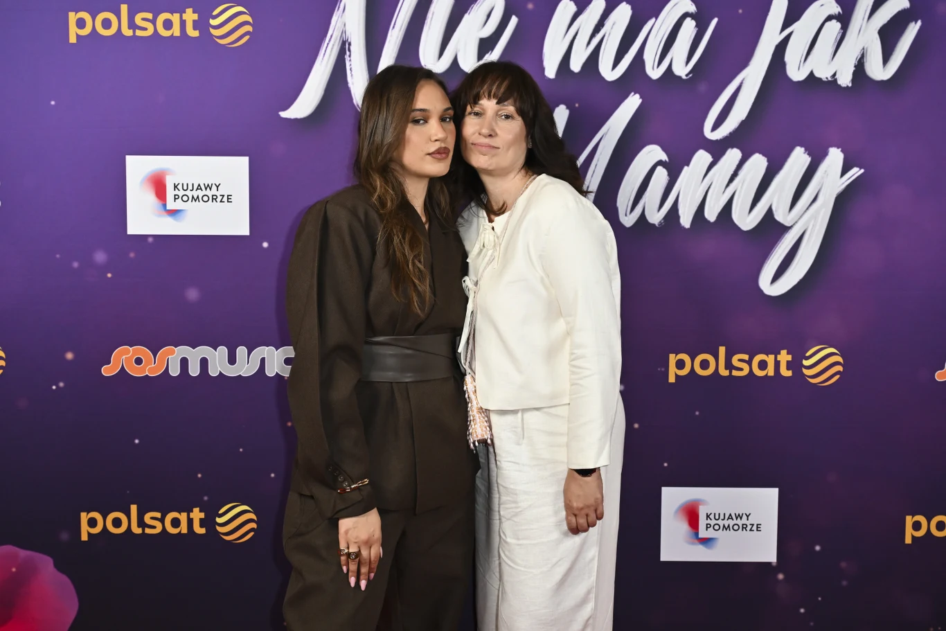 Dwie kobiety stoją obok siebie na fioletowej ściance z logo Polsat i napisem „Nie ma jak mamy”, jedna ubrana w ciemny kombinezon, druga w jasny garnitur, obejmujące się delikatnie.