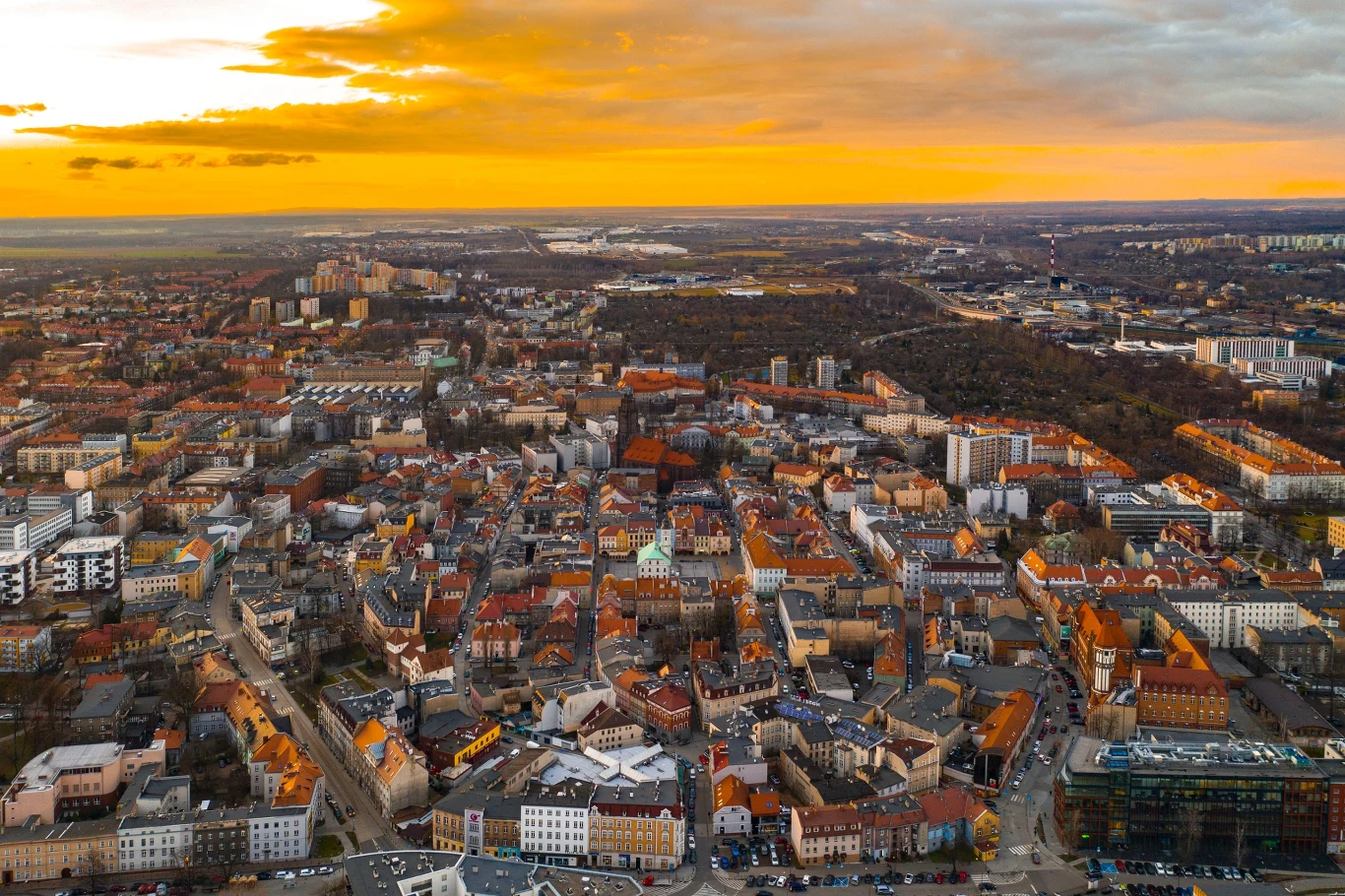 Panorama miejskiej zabudowy z dominującymi kamienicami i budynkami o czerwonych dachach, nad którymi rozciąga się żółto-pomarańczowe niebo o zachodzie słońca.