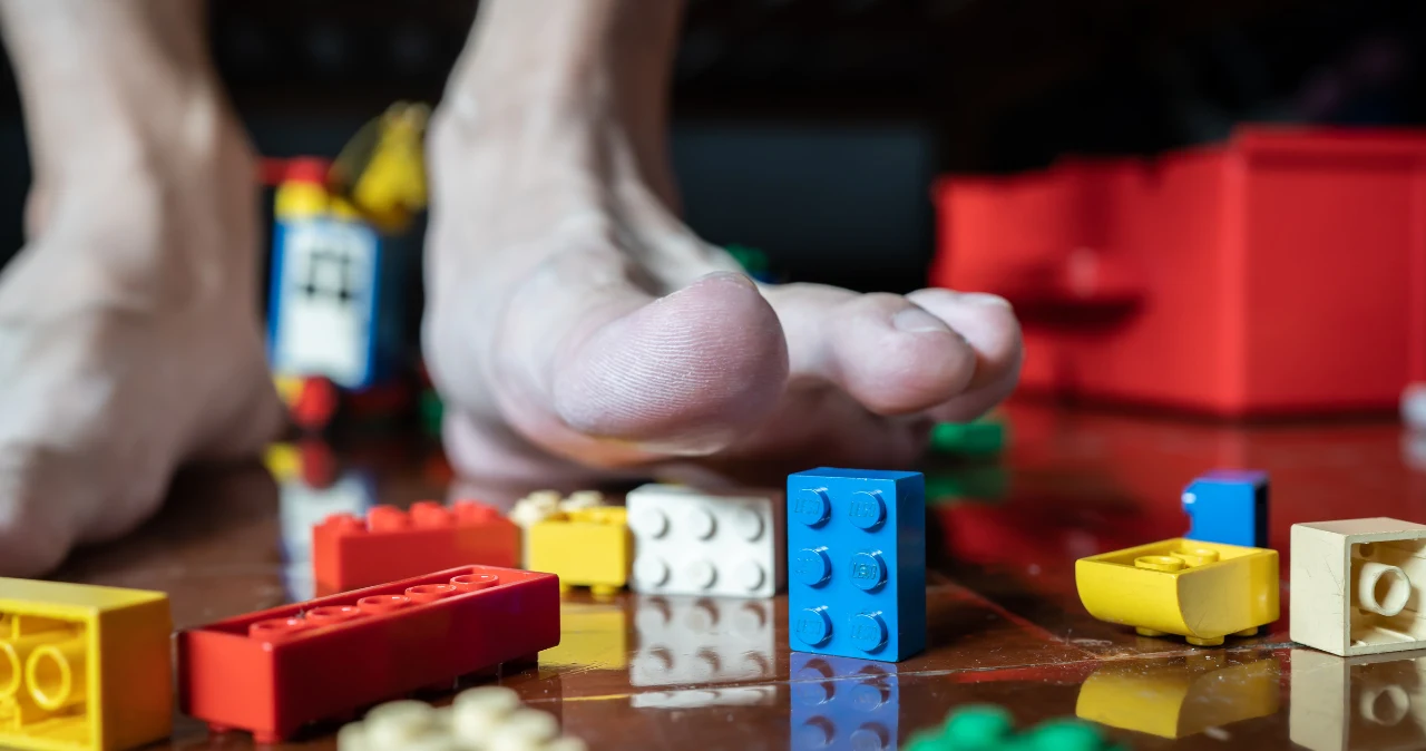 LEGO za 29,99 zł w dyskoncie! Nie spodziewałam się takich cen przed Dniem Dziecka Goła stopa przy rozsypanych klockach LEGO na drewnianej podłodze, widoczny czerwony pojemnik na zabawki.
