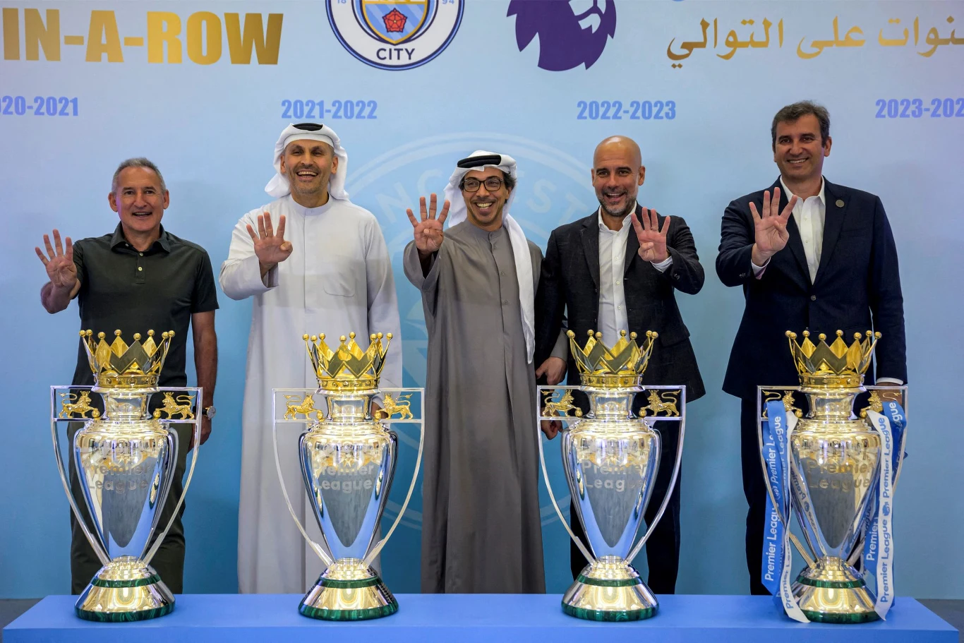 Właścicielem klubu Manchester City jest wiceprezydent ZEA Mansour bin Zayed al-Nahyan (w środku) Pięciu mężczyzn stojących za stołem z czterema pucharami Premier League, wszyscy pokazują podniesione dłonie z czterema palcami, na tle niebieskiej ścianki z napisami i logotypami klubu Manchester City oraz latami zdobycia tytułu.