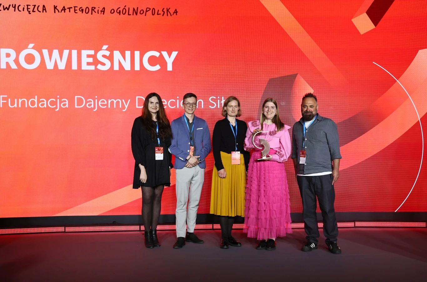 Pięć osób stojących w rzędzie na tle czerwonej ścianki z dużym napisem, jedna z nich trzyma statuetkę, wszyscy noszą identyfikatory na szyi.