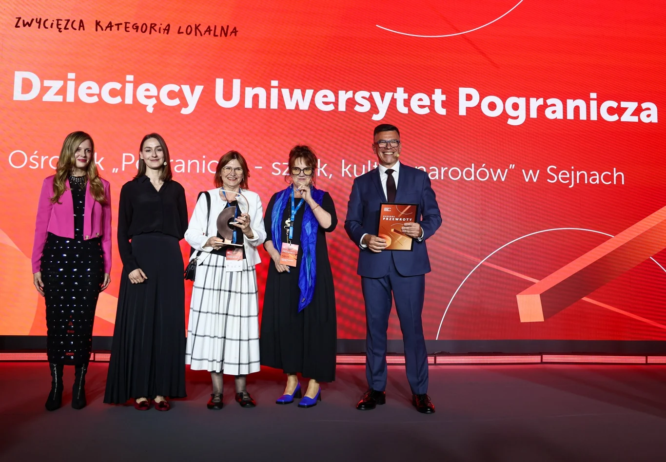 Pięć osób stojących na tle dużego, czerwonego ekranu z napisem „Dziecięcy Uniwersytet Pogranicza”, trzy kobiety trzymające statuetki, mężczyzna w garniturze trzymający dyplom, wszyscy uśmiechnięci, oficjalna atmosfera gali wręczenia nagród.