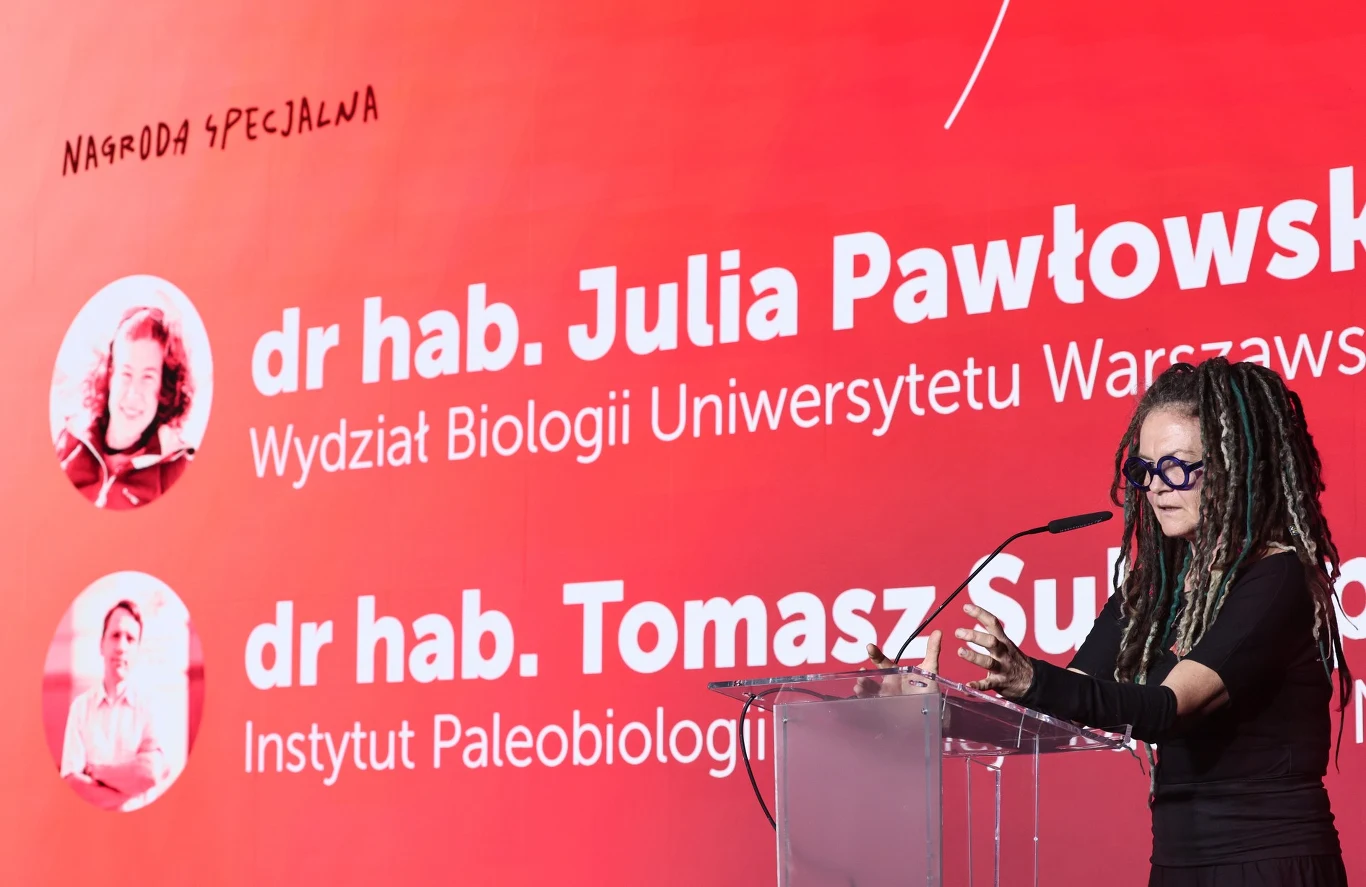 Kobieta z długimi dredami i okularami przemawia przy przezroczystej mównicy na tle czerwonej ścianki z dużymi napisami zawierającymi nazwiska i tytuły naukowe laureatów specjalnej nagrody.