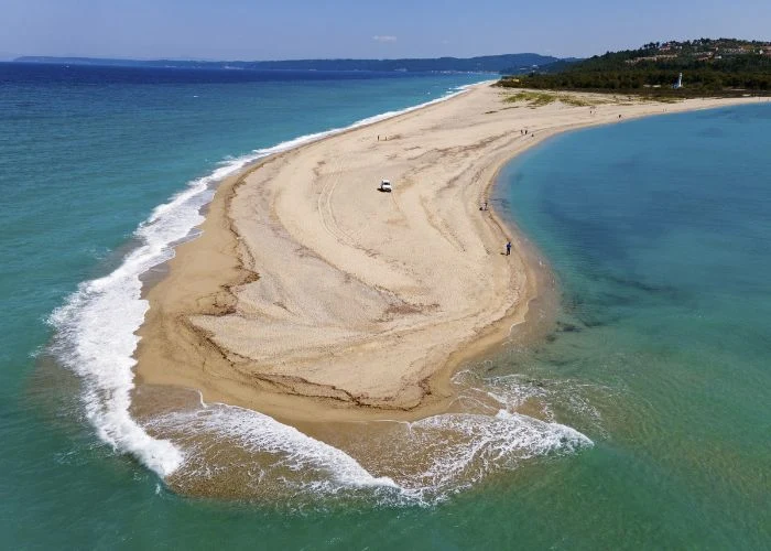 To jedna z najciekawszych plaż w całej Grecji Piaszczysty cypel plaży Possidi otoczony turkusową wodą, kilka osób spaceruje, widoczne auto terenowe, wzgórza w tle.