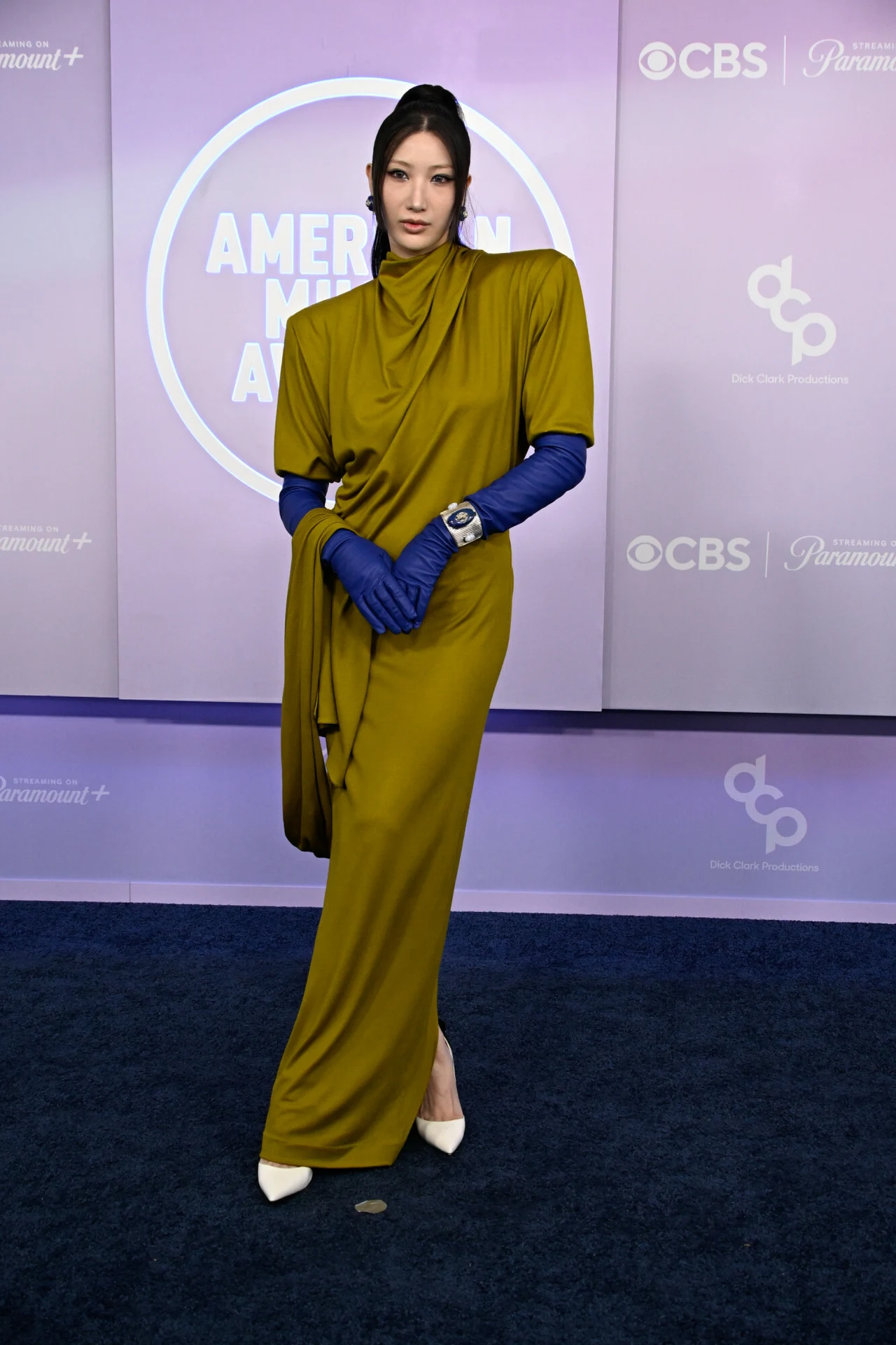 Kobieta w długiej, oliwkowej sukni z asymetrycznym krojem, uzupełnionej niebieskimi rękawiczkami i białymi szpilkami, pozuje na tle z logo gali American Music Awards.