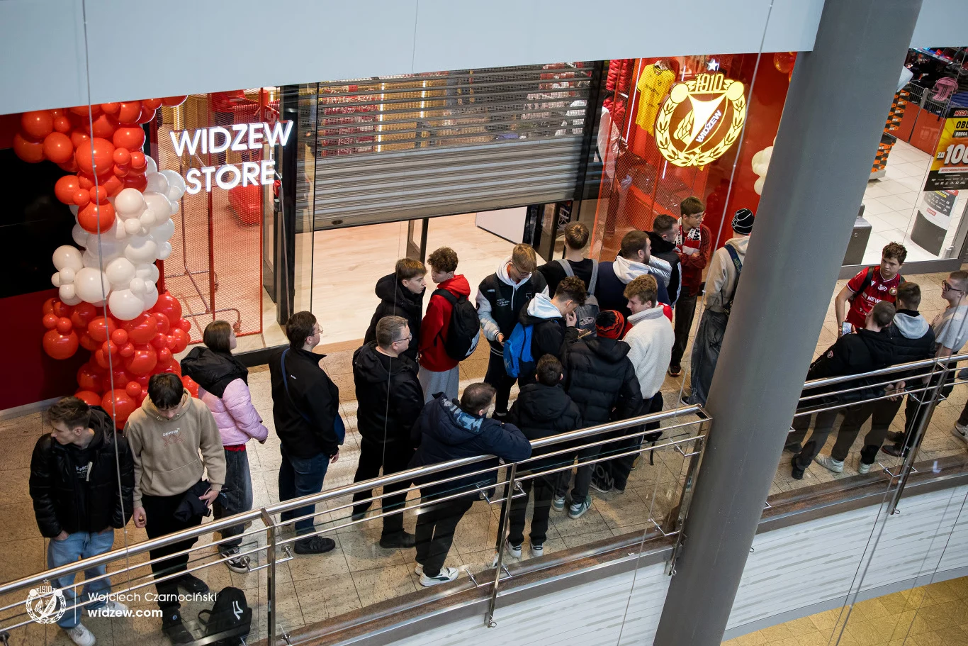 Grupa osób ustawiona w kolejce przed zamkniętym jeszcze sklepem Widzew Store, przy wejściu udekorowanym czerwono-białymi balonami.