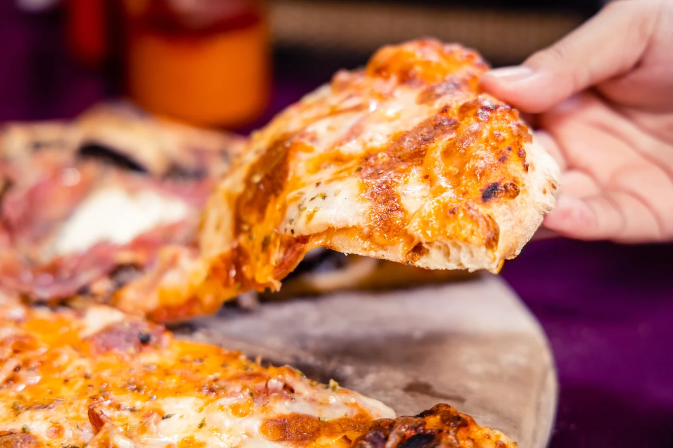 Kawałek pizzy z ciągnącym się serem podnoszony ręką z całej, upieczonej na kamieniu pizzy.