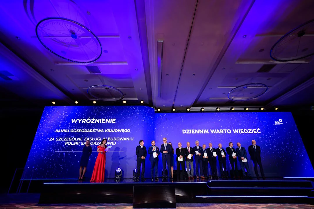Samorządy mogą ubiegać się o bezzwrotne dopłaty i kredyty w ramach prowadzonej przez BGK strategii Grupa osób elegancko ubranych stoi na scenie podczas oficjalnej gali, odbierając wyróżnienie za szczególne zasługi w budowaniu Polski, na tle niebieskiego ekranu z napisem informującym o nagrodzie.
