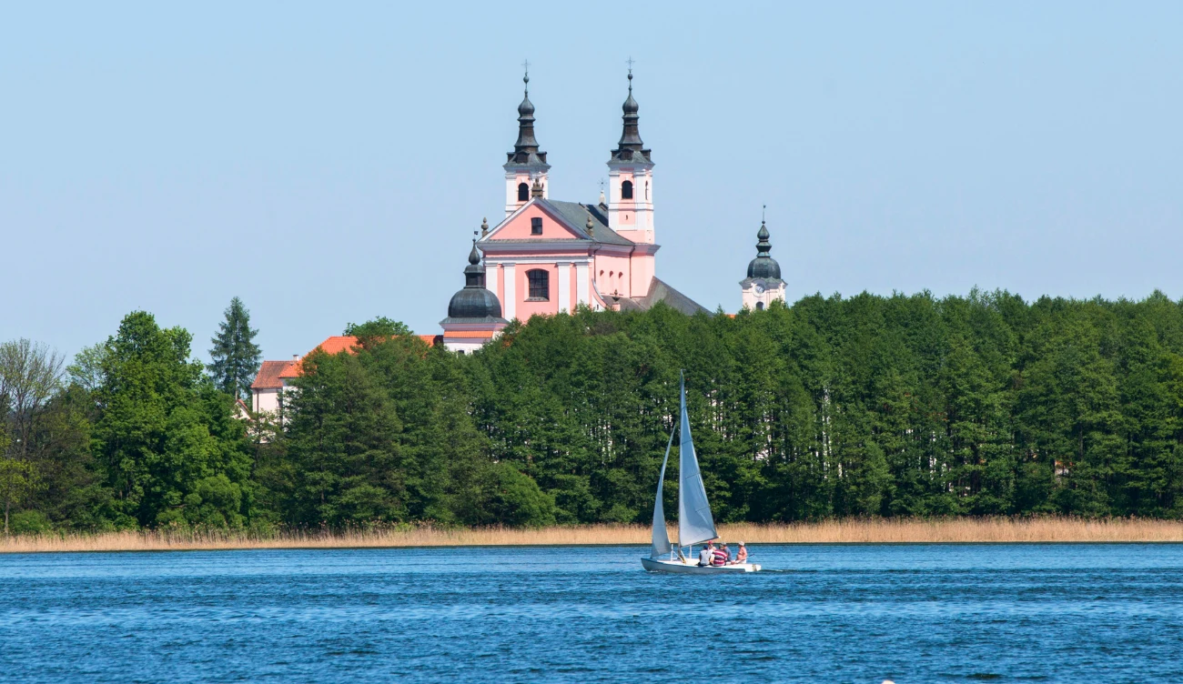 Różowy kościół z dwoma wieżami na tle zielonych drzew i niebieska żaglówka płynąca po jeziorze na pierwszym planie.