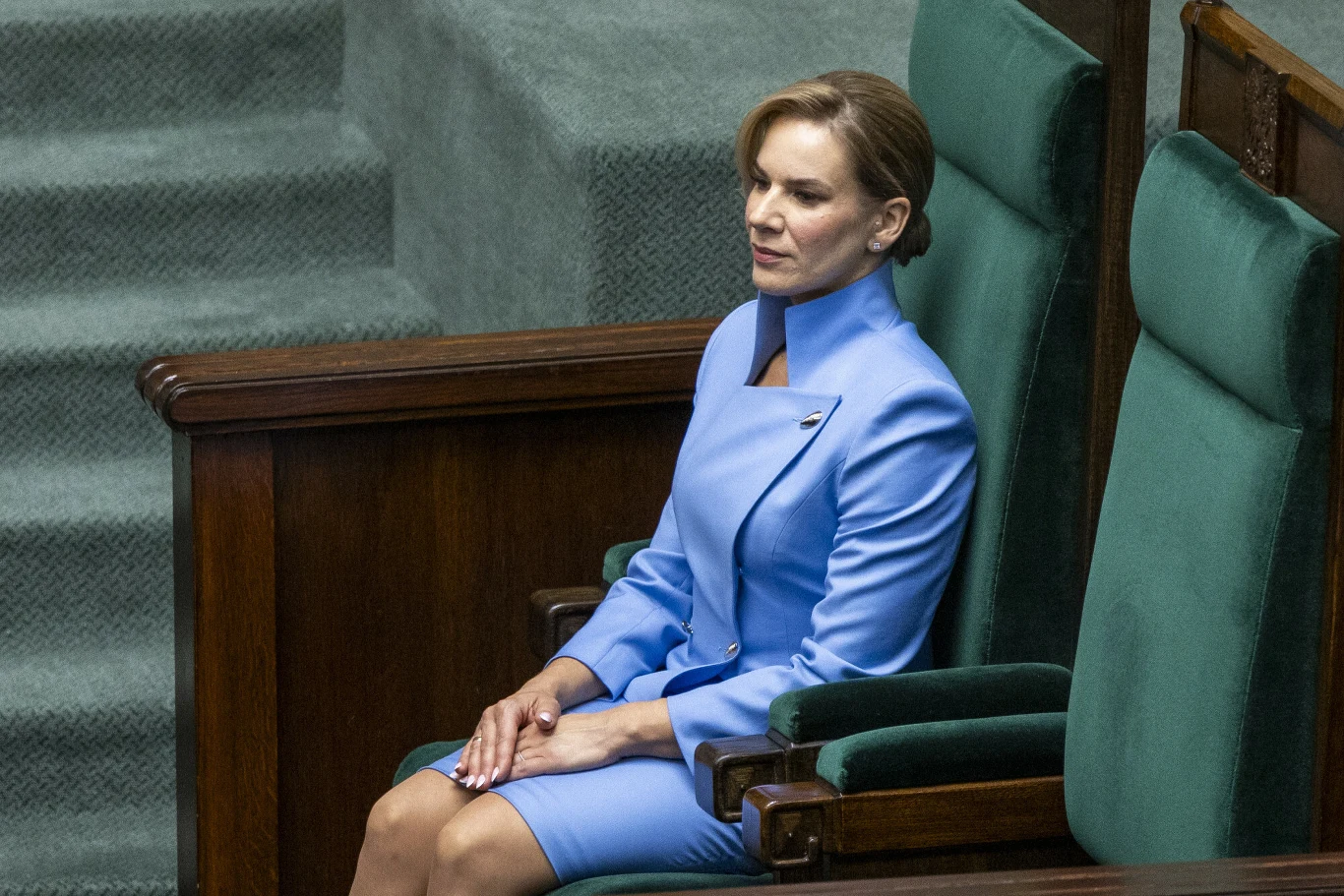 Kobieta w eleganckim, jasnoniebieskim garniturze siedzi samotnie na dużym, zielonym fotelu w przestronnym, urzędowym wnętrzu.