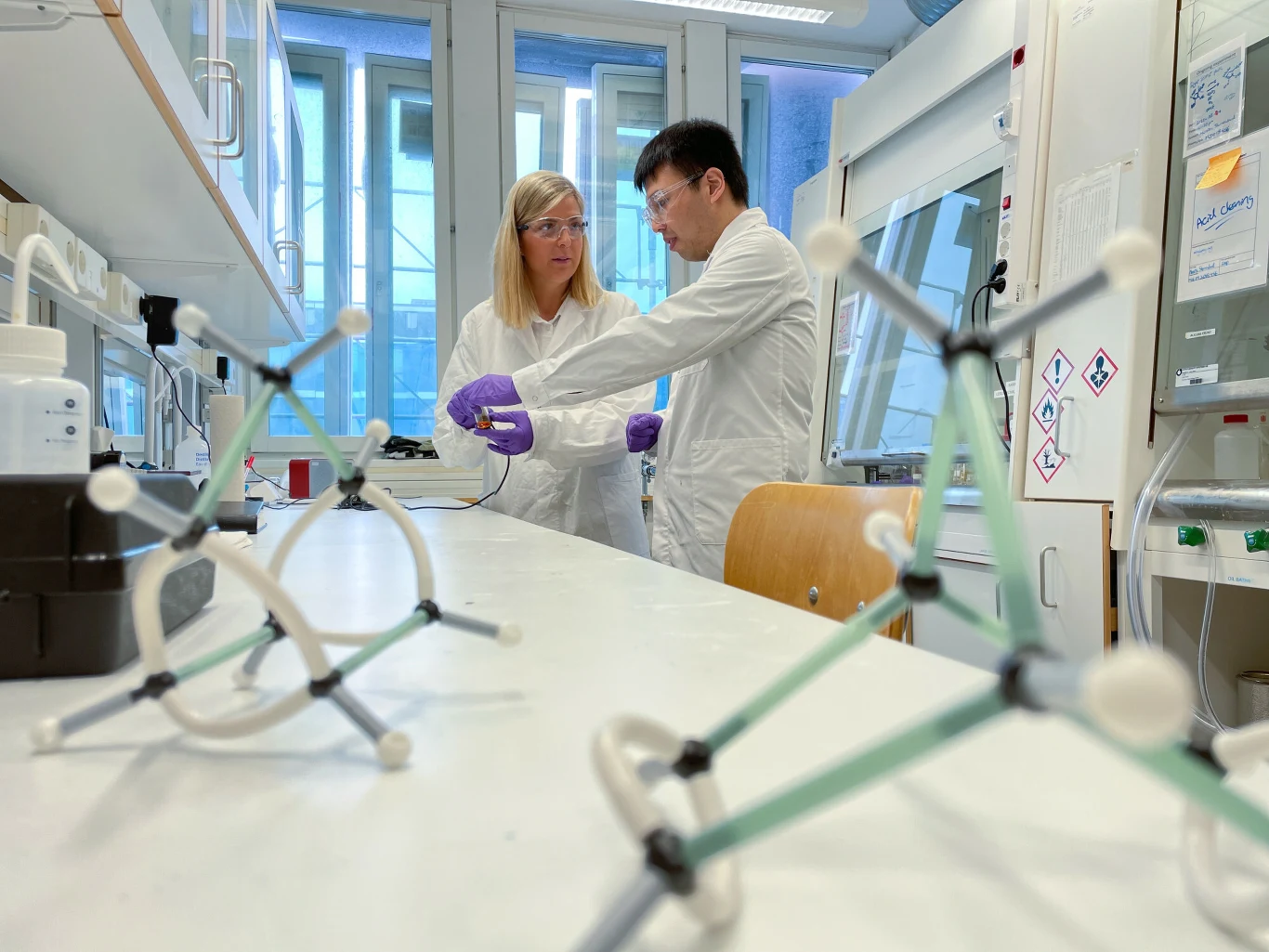 Dwoje naukowców w białych fartuchach i rękawiczkach prowadzi wspólnie badania laboratoryjne przy stole, na którym znajdują się modele cząsteczek chemicznych.
