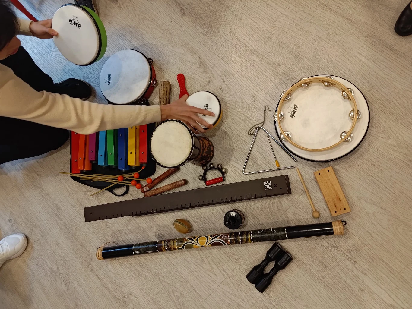 Zbiór instrumentów muzycznych obejmujący tamburyn, bębenki, kolorowy metalofon, dzwonki, drewienka, grzechotki, guiro i kij deszczowy, układanych na jasnej podłodze przez osoby widoczne po lewej stronie kadru.