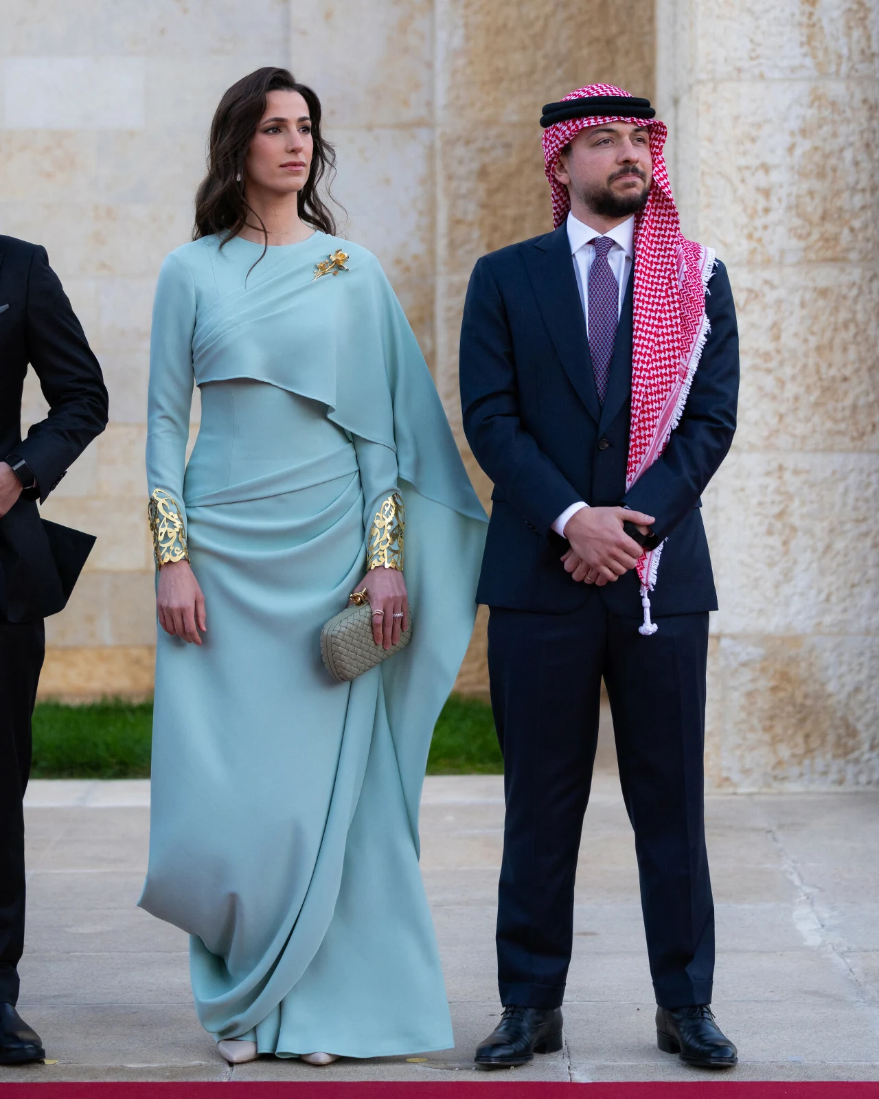Kobieta w eleganckiej, jasnoniebieskiej sukni z ozdobnymi złotymi mankietami oraz mężczyzna w granatowym garniturze i tradycyjnym arabskim nakryciu głowy stoją obok siebie na tle jasnej, kamiennej ściany.