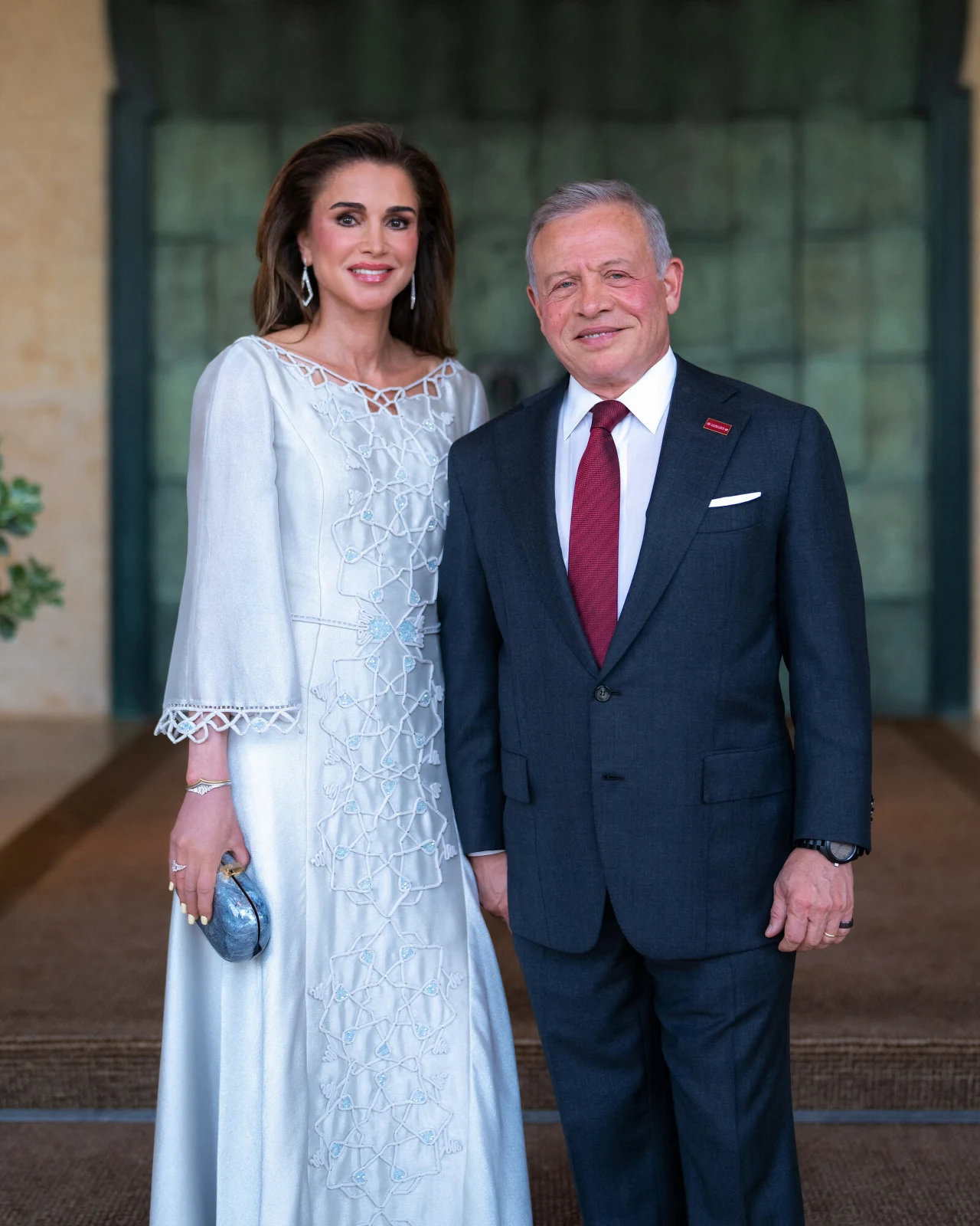 Elegancka kobieta w długiej, jasnej sukni z geometrycznymi zdobieniami stoi obok mężczyzny w ciemnym garniturze i czerwonym krawacie, oboje uśmiechnięci, gotowi do wyjścia na formalne wydarzenie.