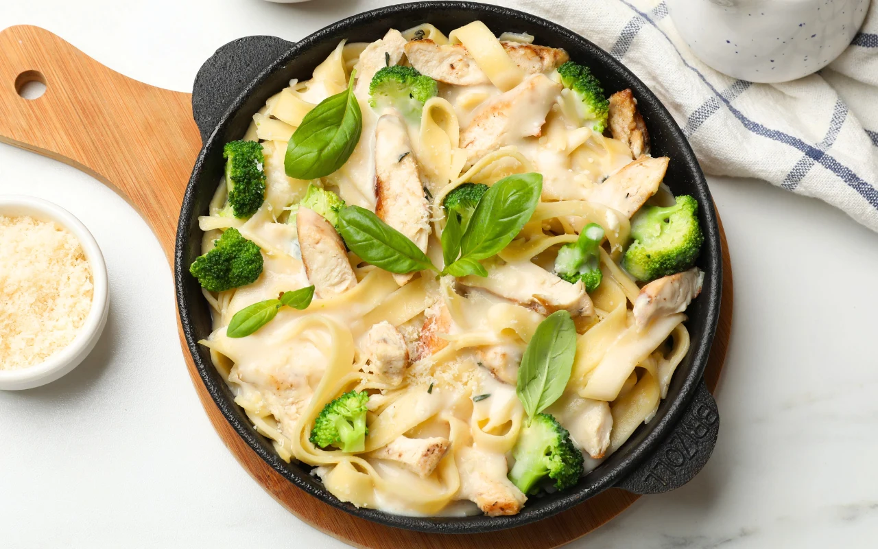 Makaron tagliatelle z kawałkami grillowanego kurczaka, różyczkami brokuła i świeżą bazylią, podany w kremowym sosie na patelni.