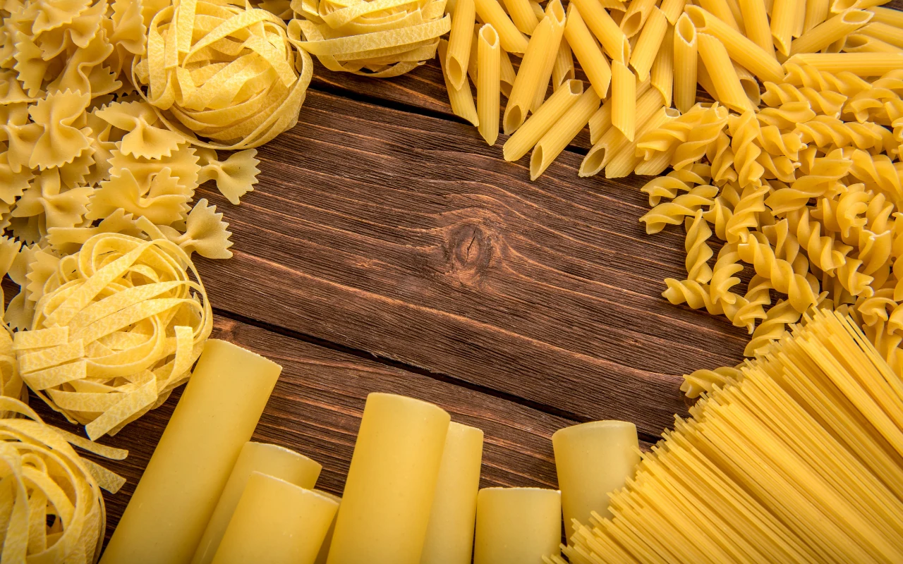 Różne rodzaje suchego makaronu, w tym penne, tagliatelle, farfalle, rigatoni, fusilli i spaghetti, ułożone wokół pustego miejsca na drewnianym stole.