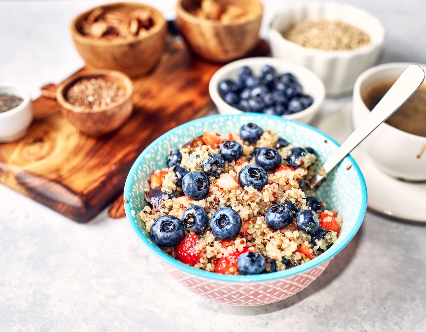 Miska kaszy quinoa z borówkami i kawałkami truskawek, obok filiżanka kawy oraz miseczki z orzechami i nasionami.
