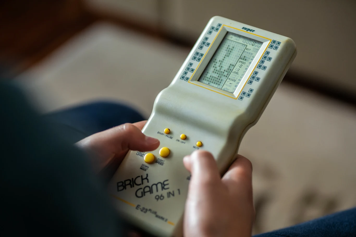 Ręce osoby trzymającej klasyczną, przenośną konsolę Brick Game z widocznym na ekranie uruchomionym Tetrisem oraz żółte przyciski sterujące na obudowie.