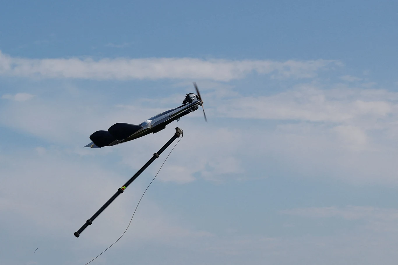 Dron z rozłożonym śmigłem unoszący się w powietrzu, podczepiony do długiego wysięgnika na tle niebieskiego, lekko zachmurzonego nieba.