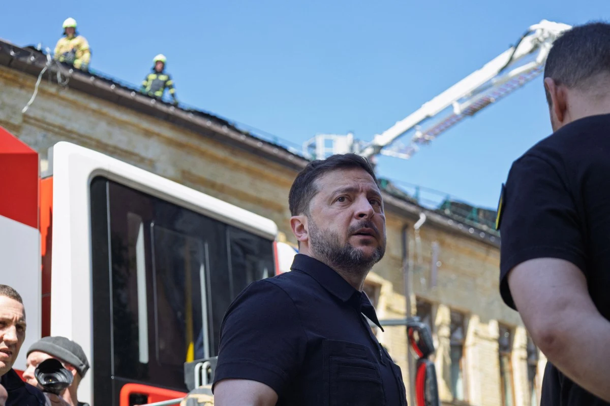 Mężczyzna w ciemnej koszulce stoi na tle strażackiego pojazdu z wysięgnikiem, obok budynku, na którego dachu pracują strażacy.