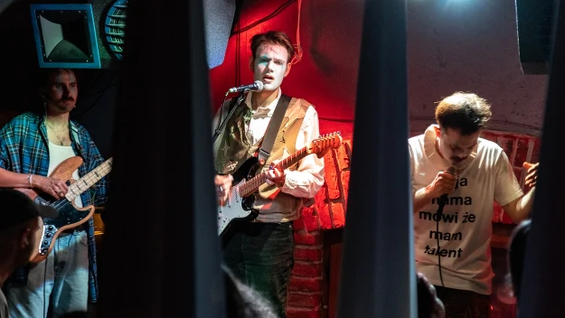 Trzech muzyków wykonujących utwór na żywo w klubie, centralny gitarzysta podkreślony czerwonym światłem, wokalista w białej koszulce z napisem oraz basista w niebieskiej koszuli po lewej stronie sceny.