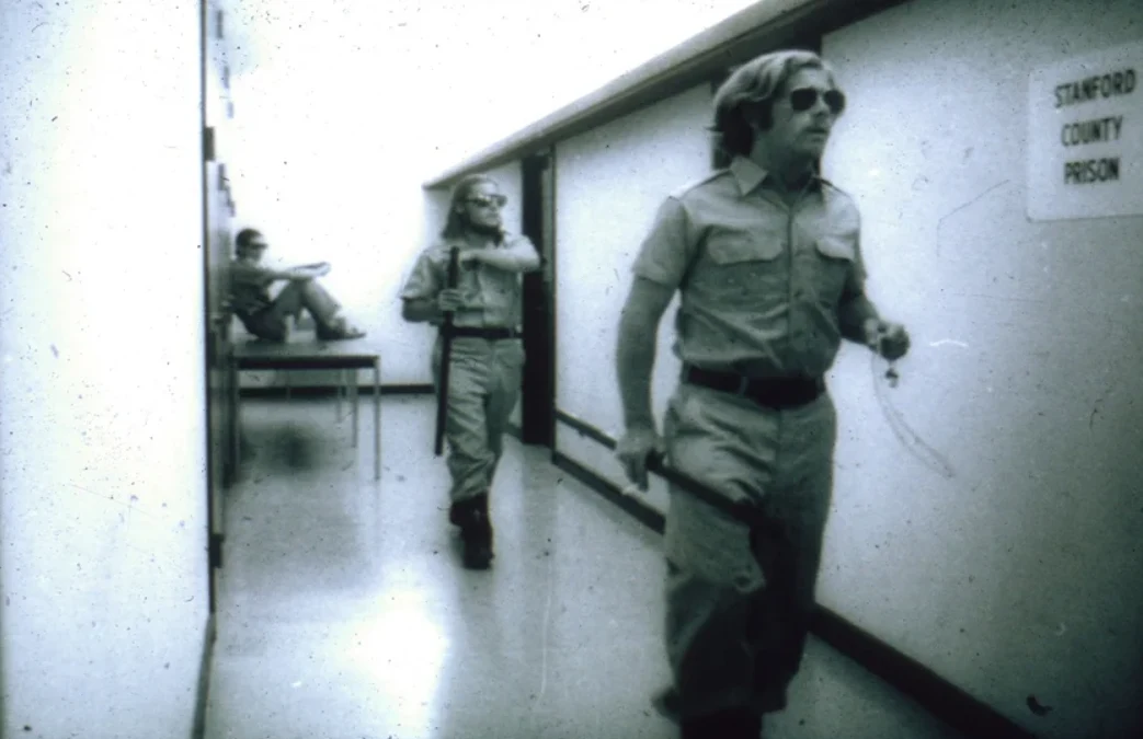 Trzech mężczyzn w mundurach wojskowych, dwóch idzie korytarzem, jeden siedzi na stole w tle; na ścianie widoczna tabliczka z napisem 'STANFORD COUNTY PRISON'.