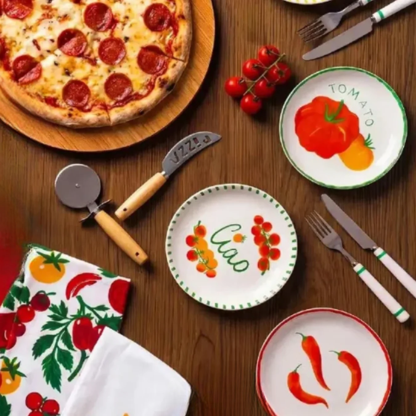 Pizza pepperoni na drewnianej desce, kolorowe talerze z motywami warzyw i napisami, zestaw sztućców oraz zestaw kuchennych tekstyliów ozdobionych wzorami pomidorów.