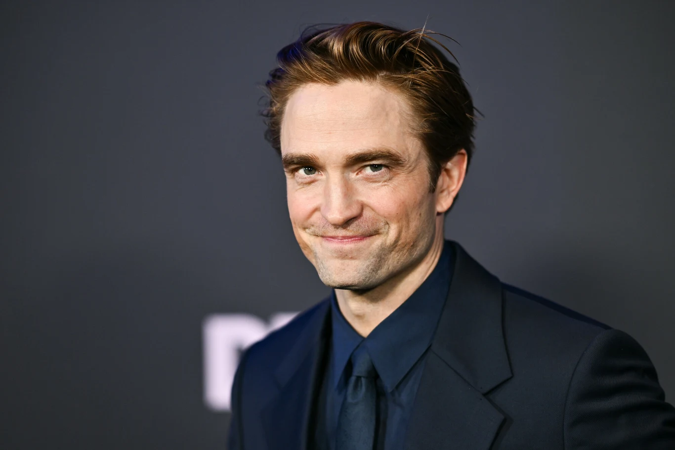 Robert Pattinson Robert Pattinson w granatowym garniturze stoi na tle ciemnej ściany z rozmytym napisem.