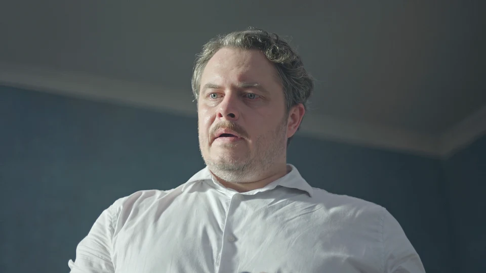 Kadr z filmu "To się nie dzieje" Mężczyzna o poważnym wyrazie twarzy ubrany w białą koszulę stoi w jasnym pomieszczeniu.