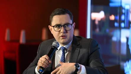 Mężczyzna w garniturze i okularach mówi do mikrofonu, siedząc przy stole na tle nowoczesnego, rozświetlonego wnętrza.