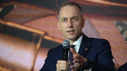 Mężczyzna w garniturze trzyma mikrofon i gestykuluje podczas wystąpienia publicznego na tle rozmytego ekranu.