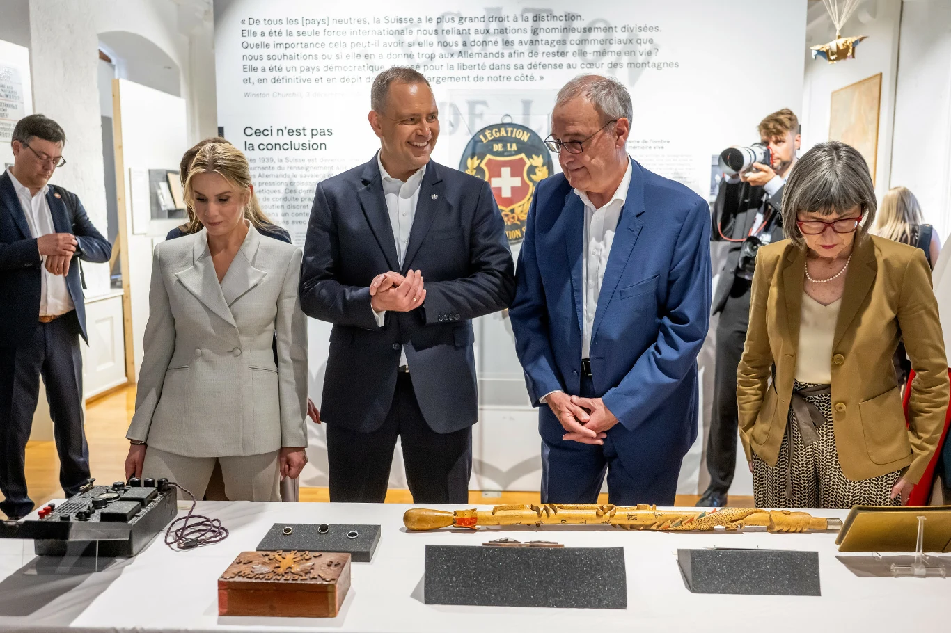 Cztery osoby w eleganckich strojach stoją przy stole, na którym leżą zabytkowe przedmioty, w tym drewniane i metalowe artefakty, zwracając na nie uwagę podczas oficjalnego wydarzenia muzealnego.