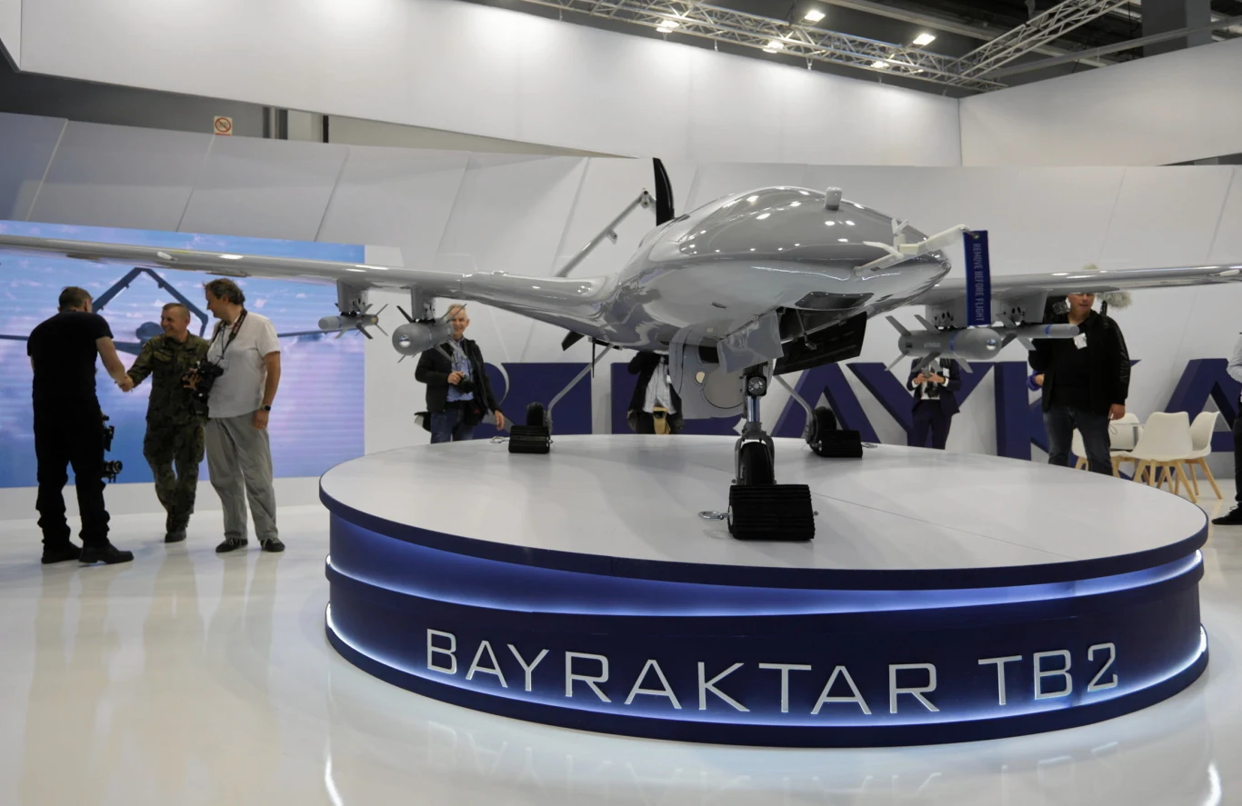 Bezzałogowy dron bojowy Bayraktar TB2 ustawiony na podświetlonej, okrągłej platformie wystawowej, otoczony przez kilka osób przyglądających się z bliska maszynie.